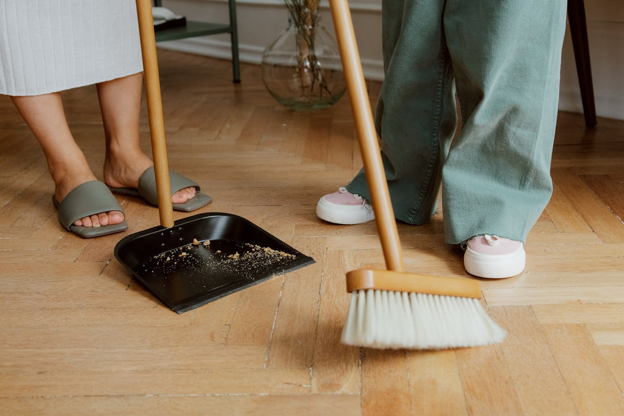 hero-img Close-up of people cleaning wooden floor with broom and dustpan indoors.
